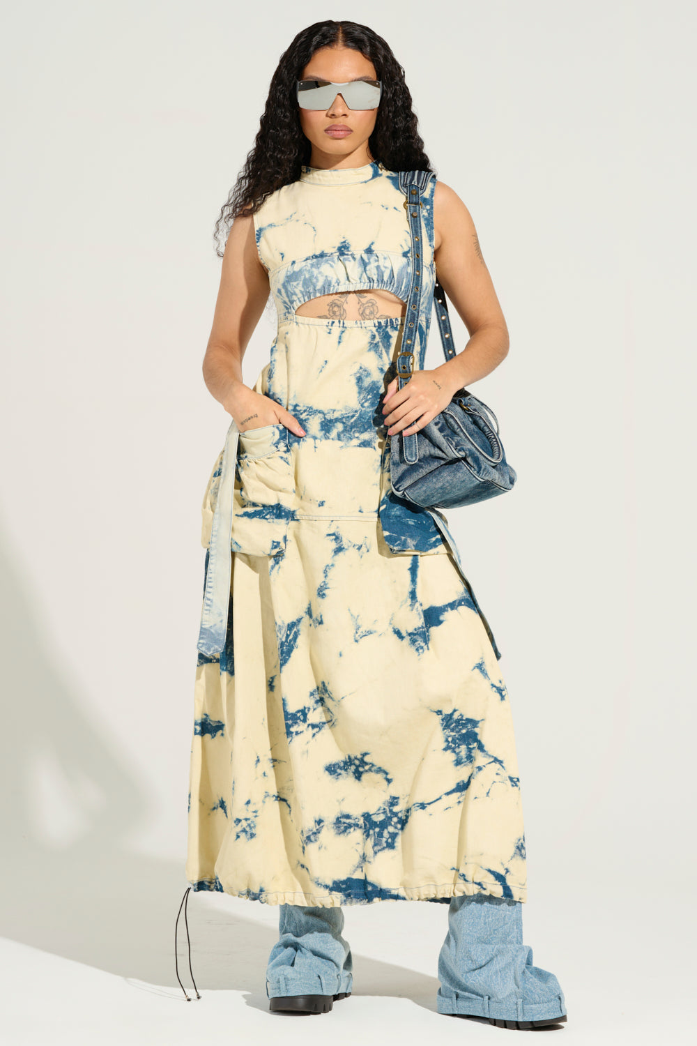 With long curly hair, a person exudes street style flair in a gorgeous STREET STYLE ACID WASH MAXI DRESS featuring blue and beige patterns with a small cutout on the front. They complete the look with large sunglasses, carry an acid wash denim handbag, and sport platform shoes.