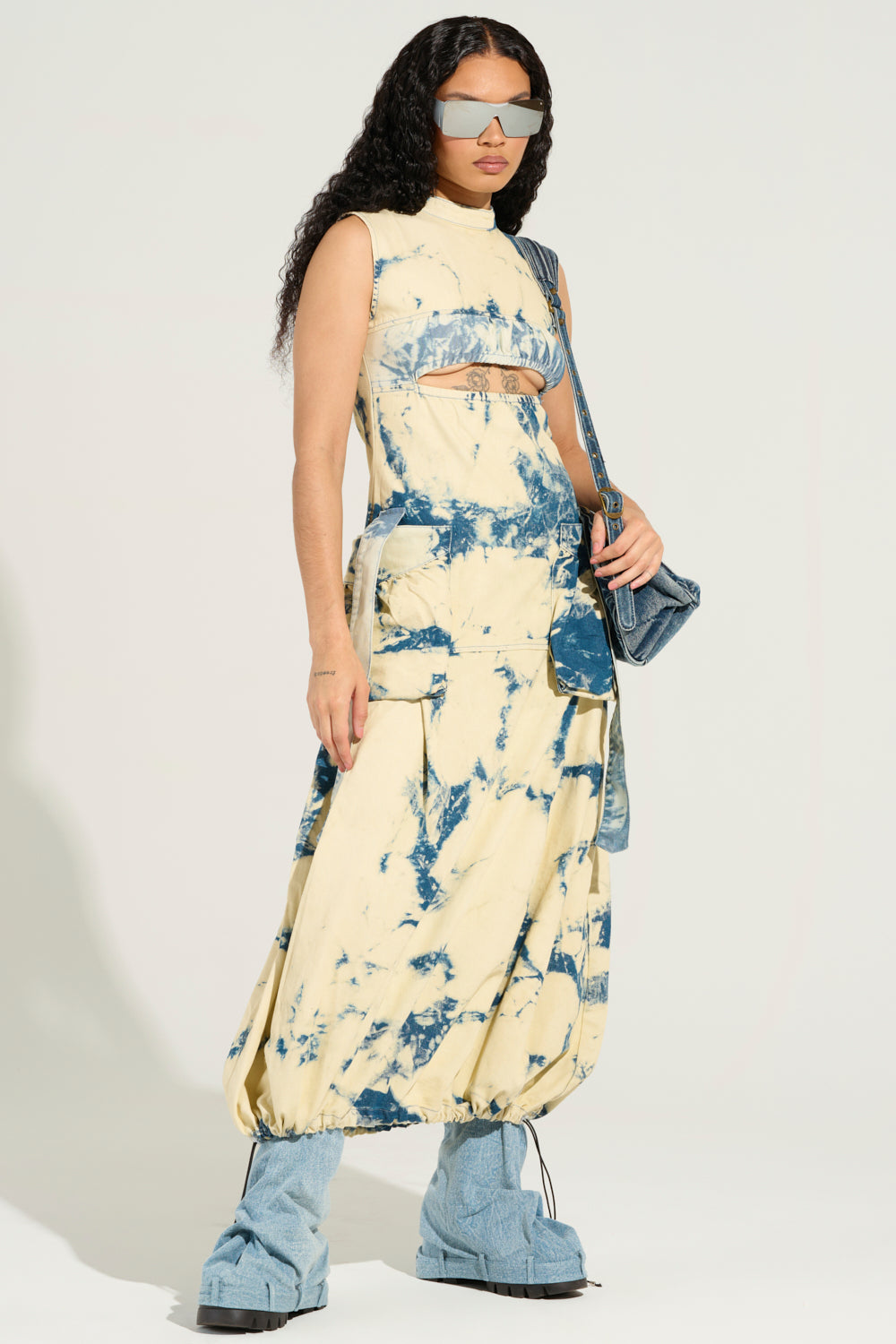 A person with long curly hair embodies classic street style, wearing reflective sunglasses and the STREET STYLE ACID WASH MAXI DRESS, complete with side pockets. They complement the dress with large blue platform shoes and carry a small blue bag, standing effortlessly cool against a plain background.