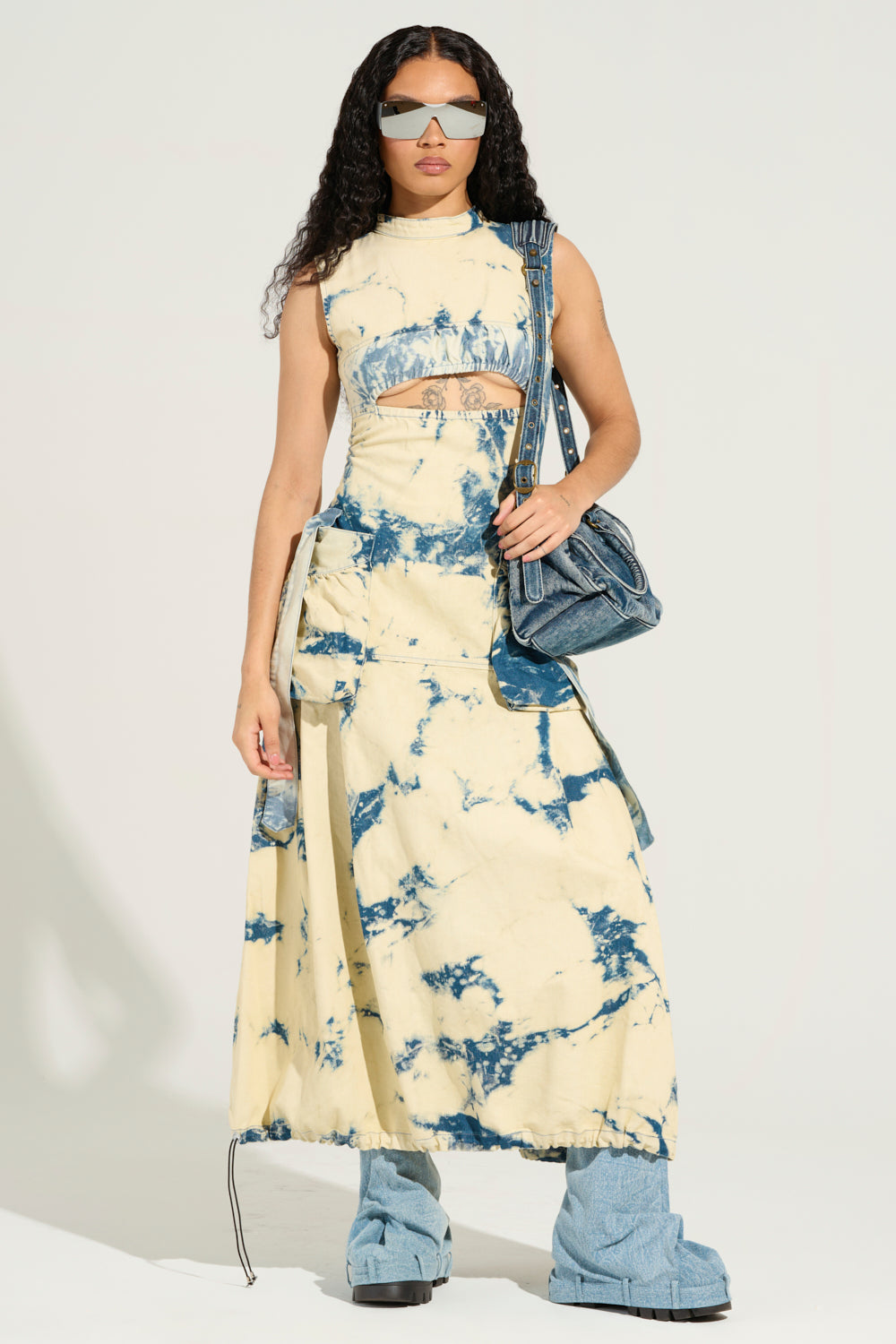A person with long curly hair wears futuristic sunglasses, a sleeveless street style acid wash maxi dress, and platform boots. They hold a denim bag over one shoulder, embodying street style confidence against a plain, light background.