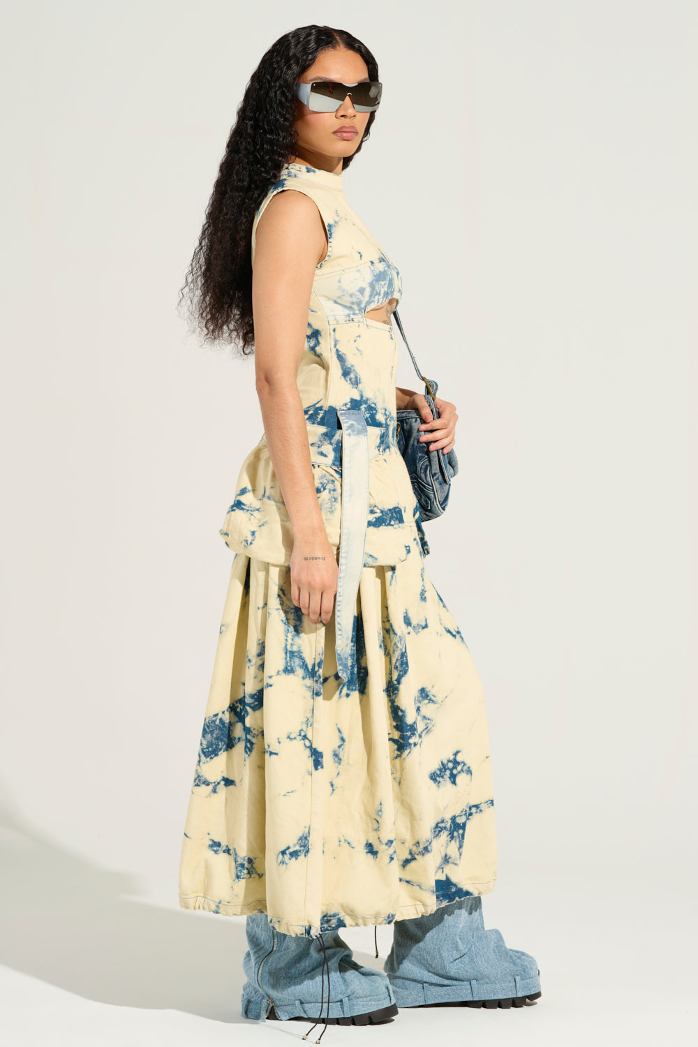 A woman stands confidently in a Street Style Acid Wash Maxi Dress, featuring hues of blue against a beige backdrop. Her long, curly hair cascades around her oversized sunglasses, while blue chunky shoes complement the look. Embracing street style with flair, she carries a matching crossbody bag for an effortlessly chic appearance.