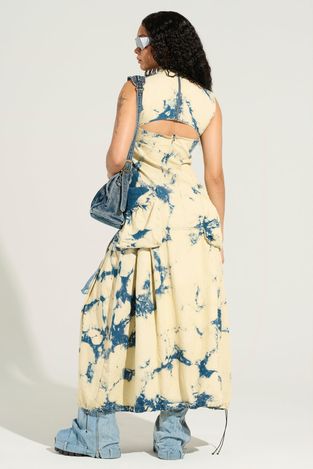 Dressed in the STREET STYLE ACID WASH MAXI DRESS, a person stands with their back to the camera, showcasing street fashion. They hold an acid wash denim bag and pair it with denim boots and sunglasses, while dark curly hair cascades down their back.