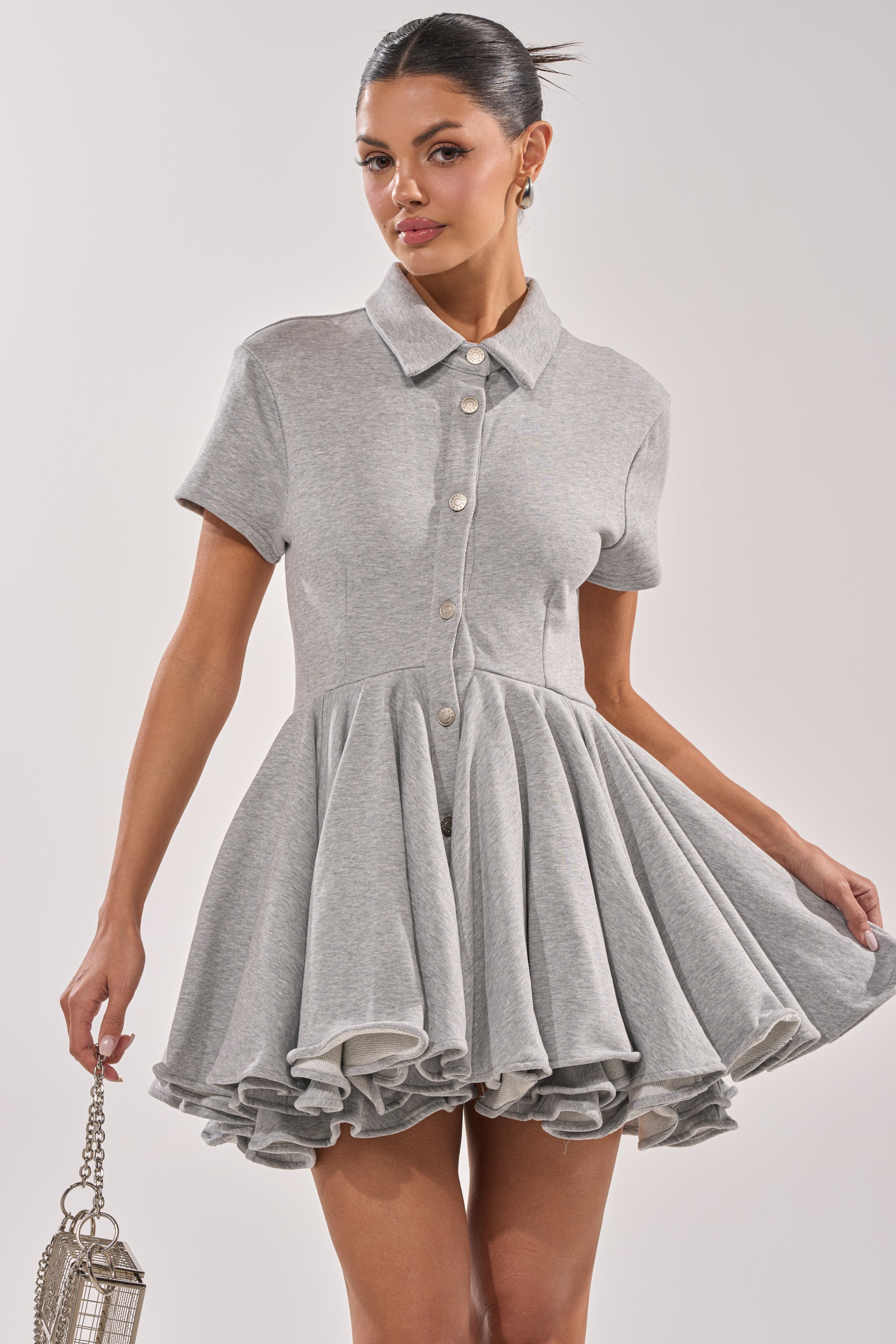She wears the AVERY SHORT SLEEVE FRENCH TERRY MINI DRESS in heather grey, featuring a collared neckline, flared ruffled skirt, and short sleeves. She completes her look with a small silver handbag and a sleek updo. 1