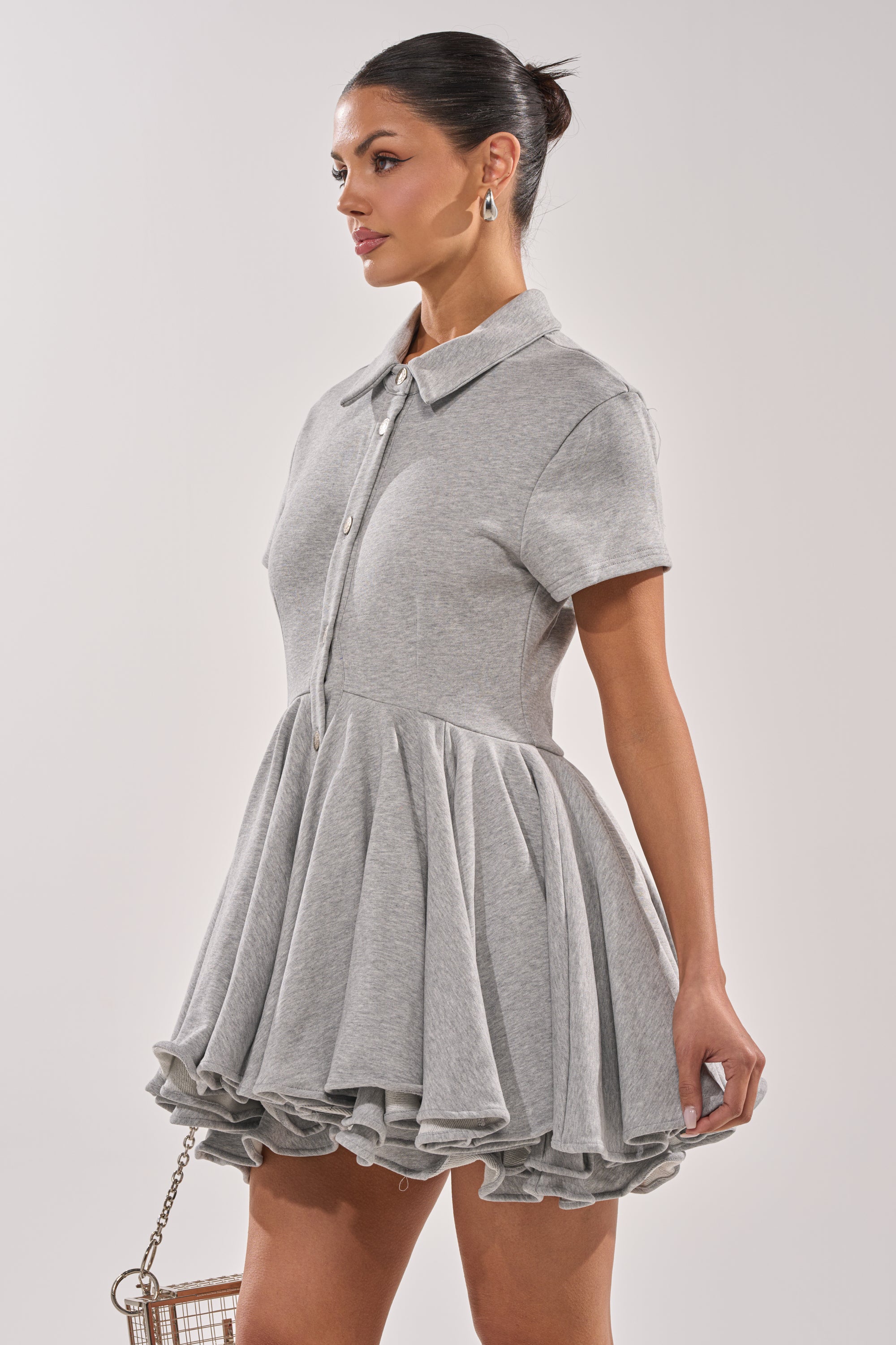 Wearing the AVERY SHORT SLEEVE FRENCH TERRY MINI DRESS in heather grey, a woman stands sideways with her hair tied back, holding a small wireframe square purse. The dress features a collared neckline, flared ruffled skirt, and button-down front. 2