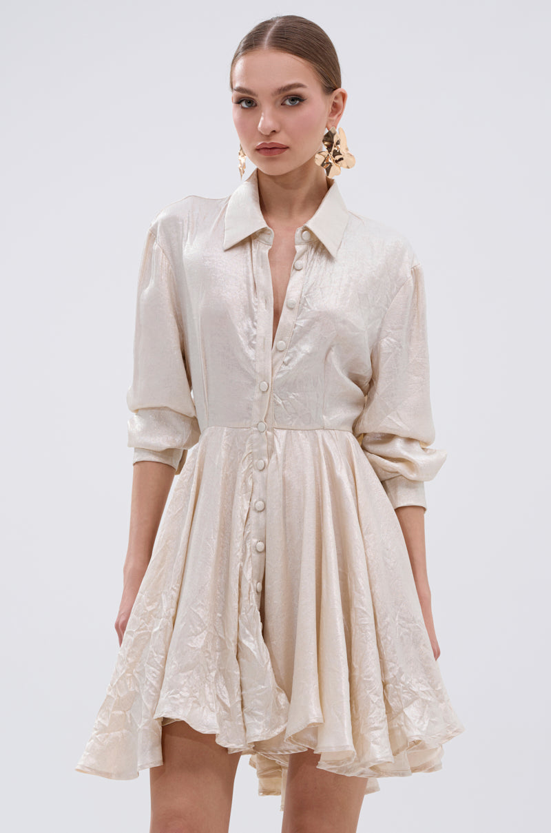 A woman poses against a plain light background in THE IT GIRL IVORY METALLIC MINI DRESS, featuring long sleeves and a flared pleated mini skirt. Her hair is tied back to highlight her statement earrings, adding glamour to the look.
