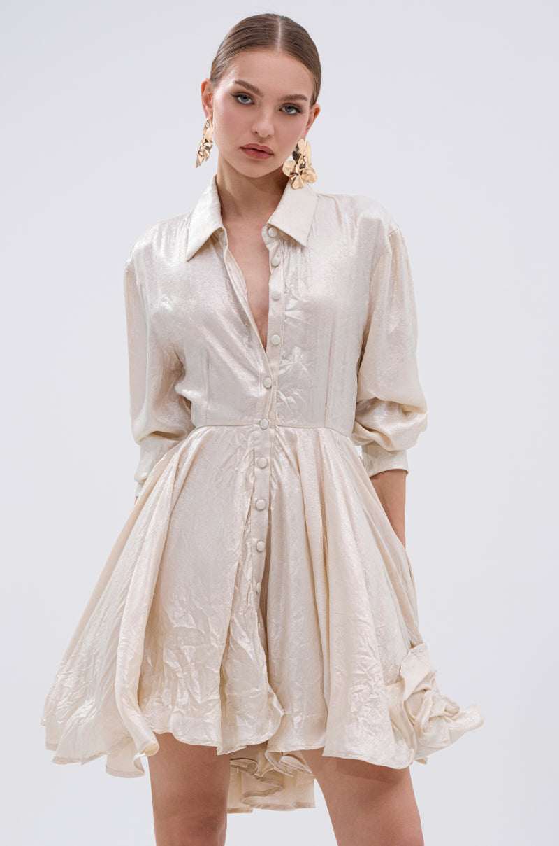 A woman wears THE IT GIRL IVORY METALLIC MINI DRESS, featuring a flared pleated skirt and rolled-up sleeves, styled with statement earrings. This glamorous piece stands out with its shimmery cream hue and effortless elegance.