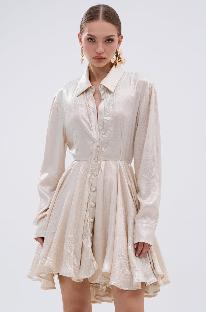 A woman wears the THE IT GIRL IVORY METALLIC MINI DRESS, featuring a collared neckline, button-down front, and flared pleated mini skirt. She pairs it with gold statement earrings while standing against a plain light background.