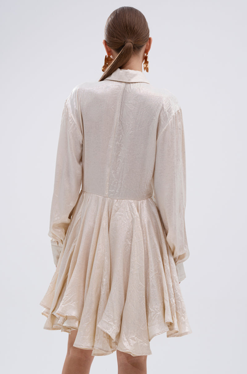 A woman with straight brown hair tied back is seen from the back wearing THE IT GIRL IVORY METALLIC MINI DRESS, featuring long sleeves and a flared pleated mini skirt, standing against a plain light background.