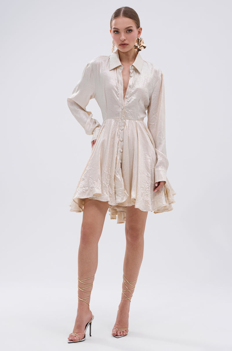A woman exudes confidence in THE IT GIRL IVORY METALLIC MINI DRESS, featuring a flared pleated mini skirt, styled with statement earrings, strappy high-heeled sandals, and her hair pulled back.