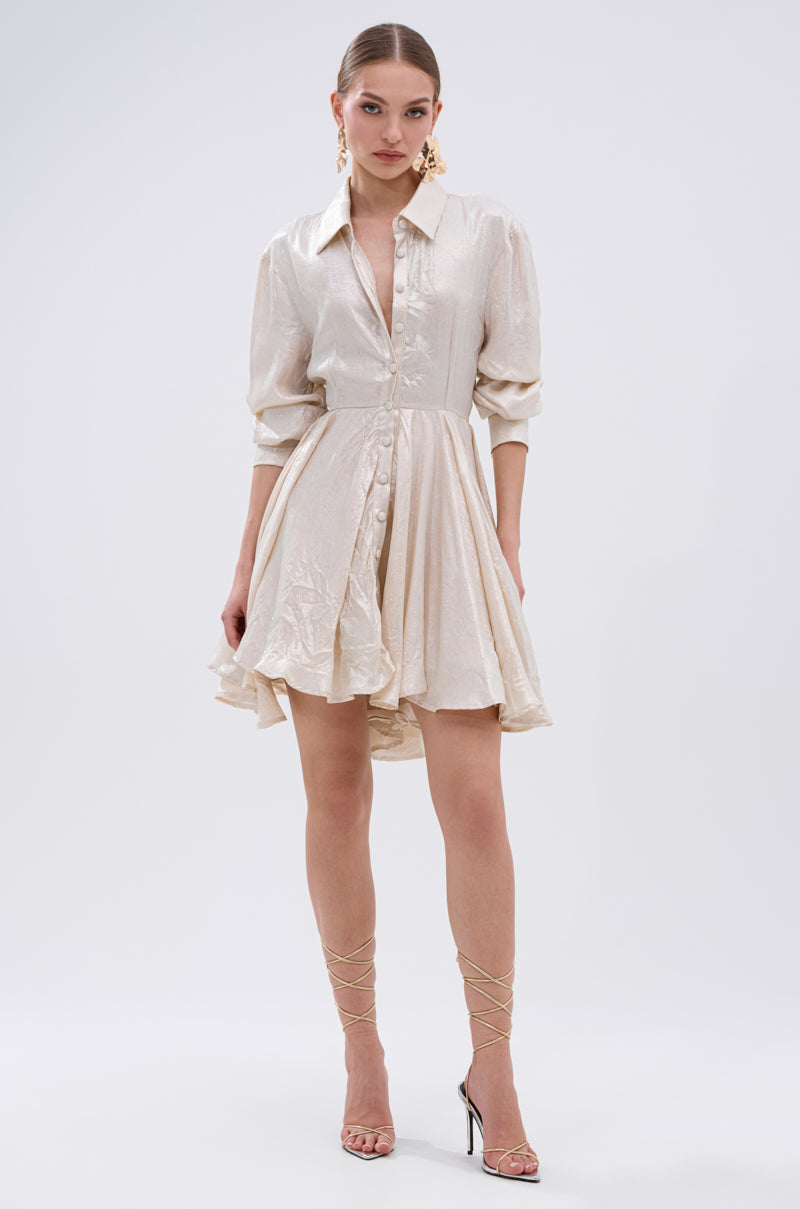 A woman stands against a plain white background wearing THE IT GIRL IVORY METALLIC MINI DRESS with long sleeves and a flared pleated mini skirt, paired with strappy high-heeled sandals and statement earrings for a glamorous look.