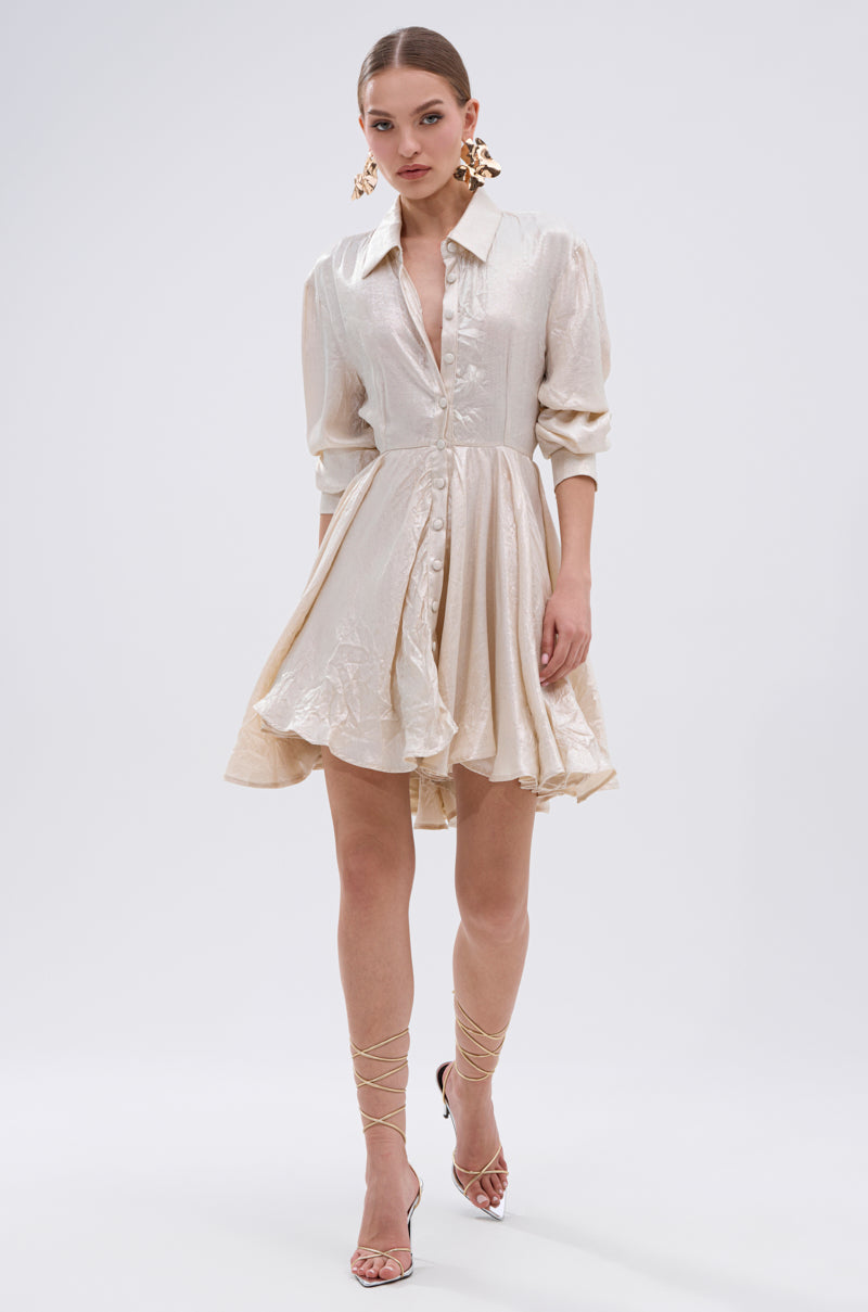 A woman poses against a plain white background in THE IT GIRL IVORY METALLIC MINI DRESS, featuring long sleeves and a flared pleated mini skirt, paired with statement earrings and strappy high heels.