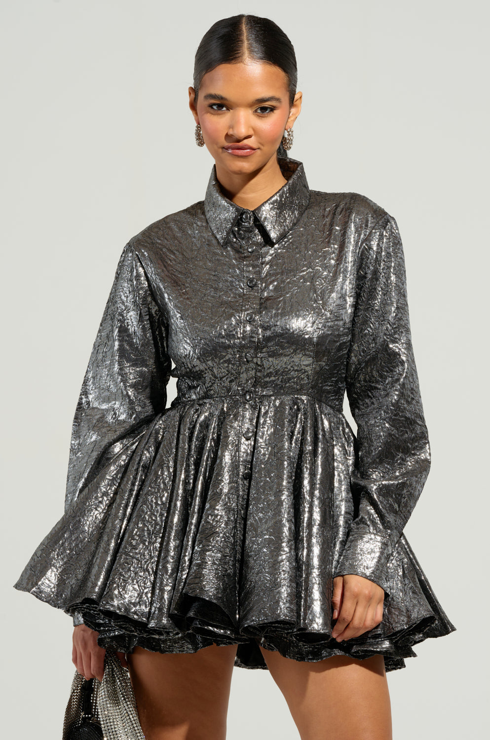 Dressed in THE IT GIRL SILVER METALLIC MINI DRESS, a figure stands against a plain background. They carry a sparkling handbag and wear hoop earrings, embodying the confidence and stylish allure of an It Girl. 2