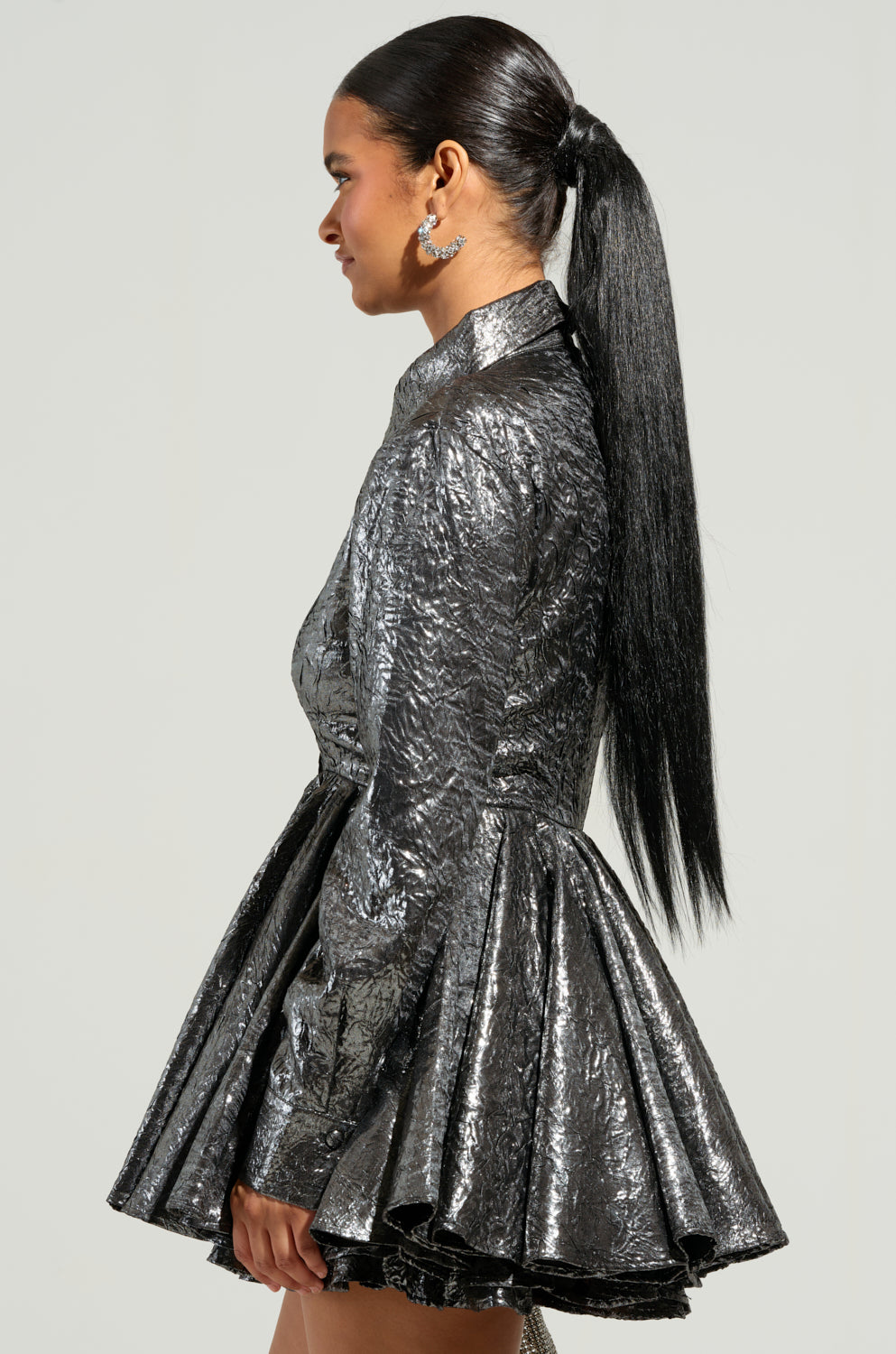 A person facing sideways is styled in THE IT GIRL SILVER METALLIC MINI DRESS, featuring a chic flared skirt and long sleeves. They exude an "It Girl" aura, with long, straight black hair in a ponytail and large, ornate earrings. The plain light-colored background accentuates their standout appearance. 3