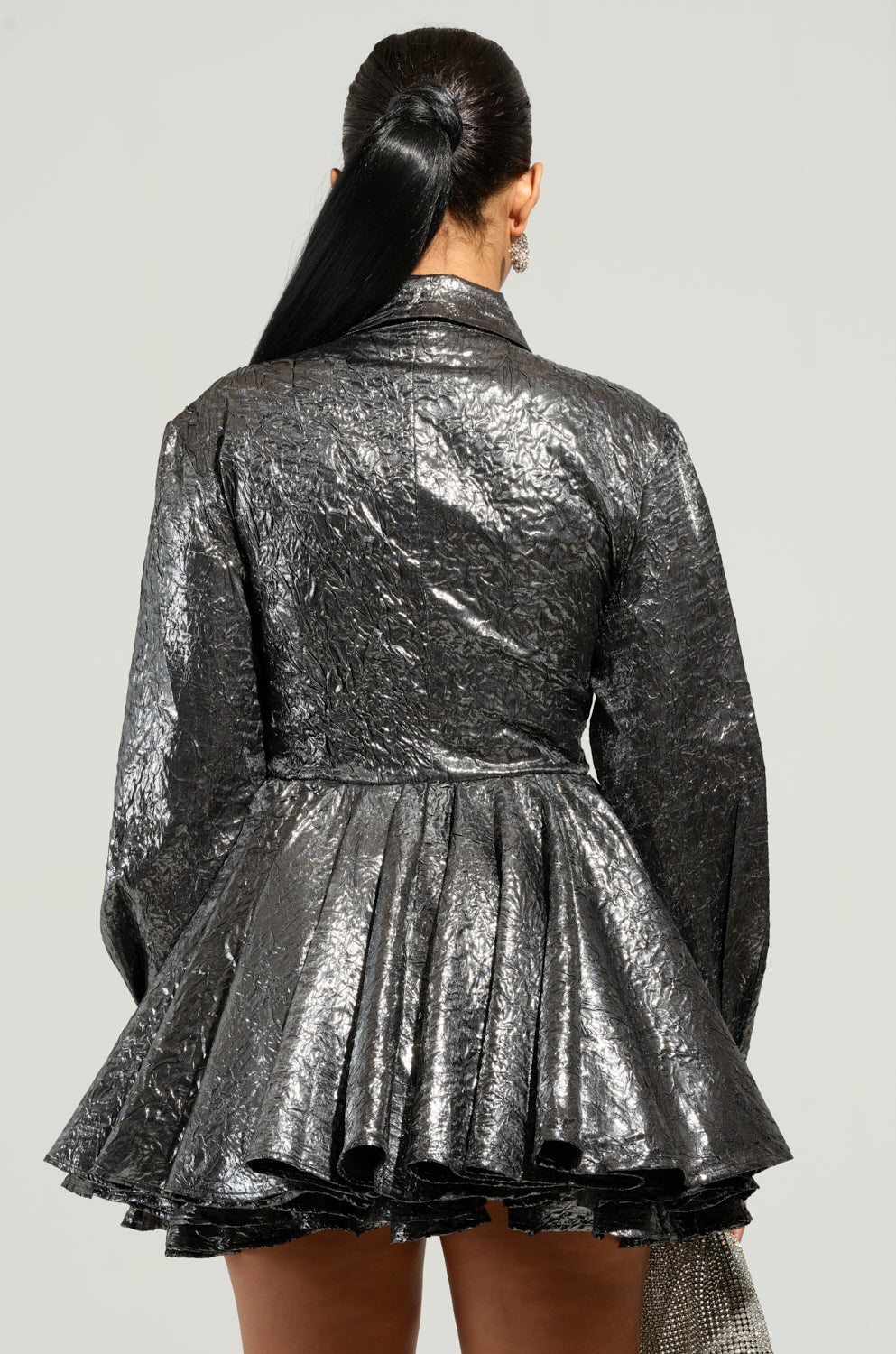 Rear view of an It Girl with long, dark hair tied in a ponytail, wearing THE IT GIRL SILVER METALLIC MINI DRESS. The plain background highlights the glossy texture of her chic ensemble. 4