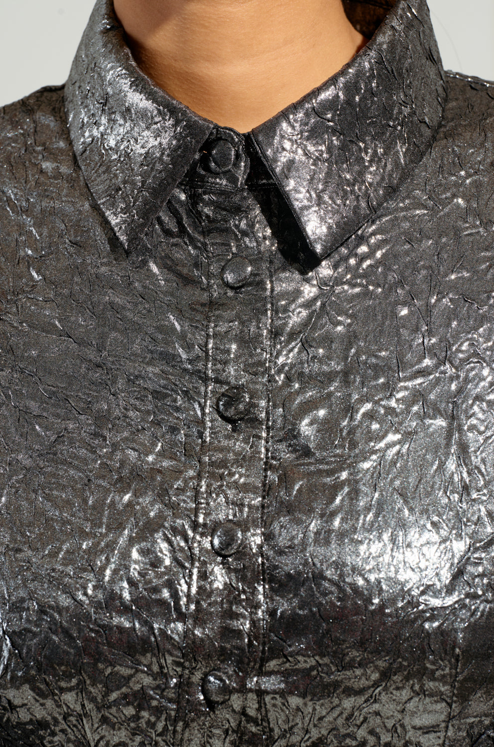 A close-up of a person wearing *THE IT GIRL SILVER METALLIC MINI DRESS*, featuring a shiny, textured metallic fabric that glistens with light to create a striking glossy effect. The pointed collar and round black buttons enhance the crinkled look, echoing the quintessential It Girl style. 6