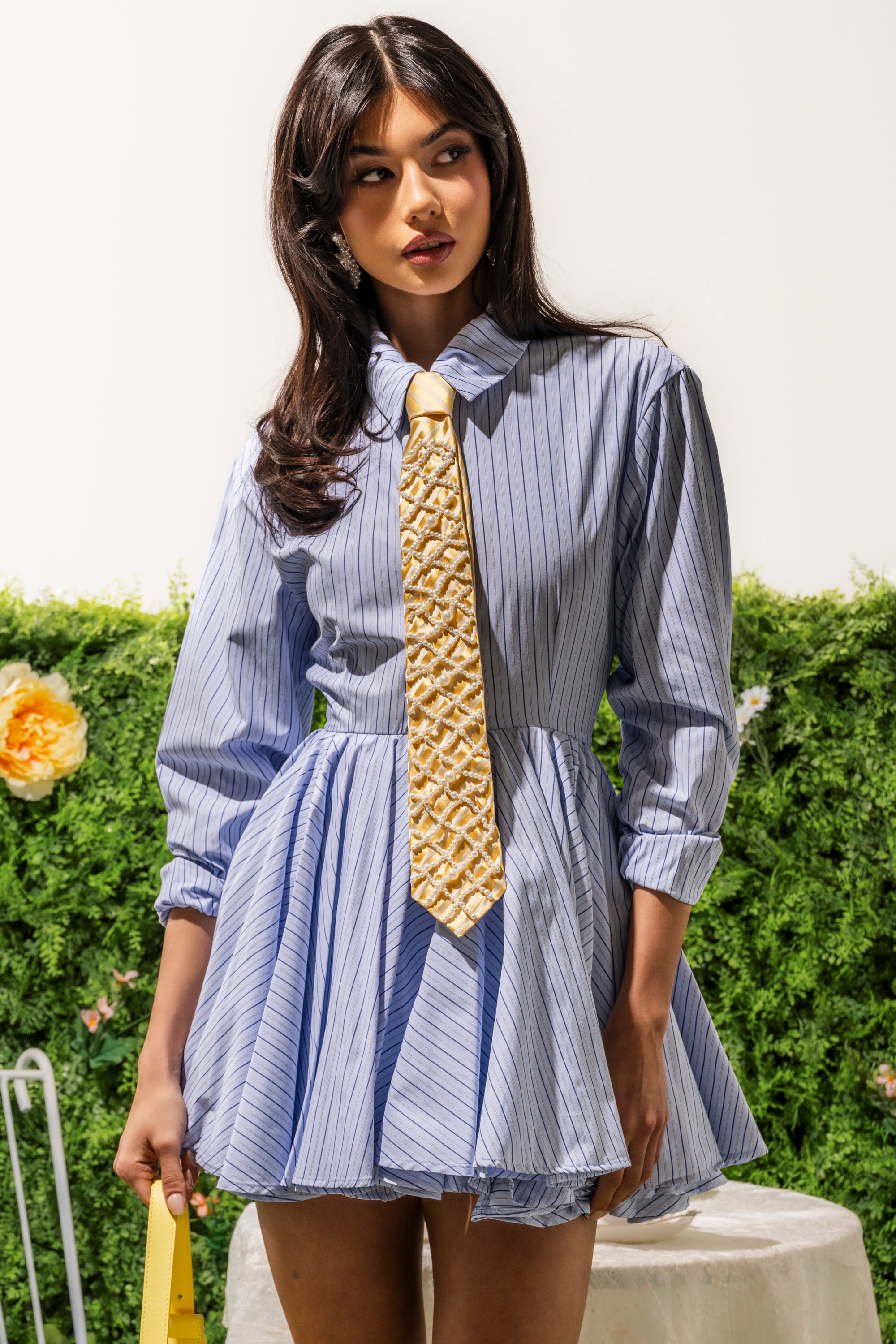 A woman stands outdoors in the HAPPY HOUR PINSTRIPE MINI DRESS, featuring a blue-and-white cotton fabric and pleated skirt. She has long dark hair, looks to the side, and is surrounded by greenery and flowers. 0