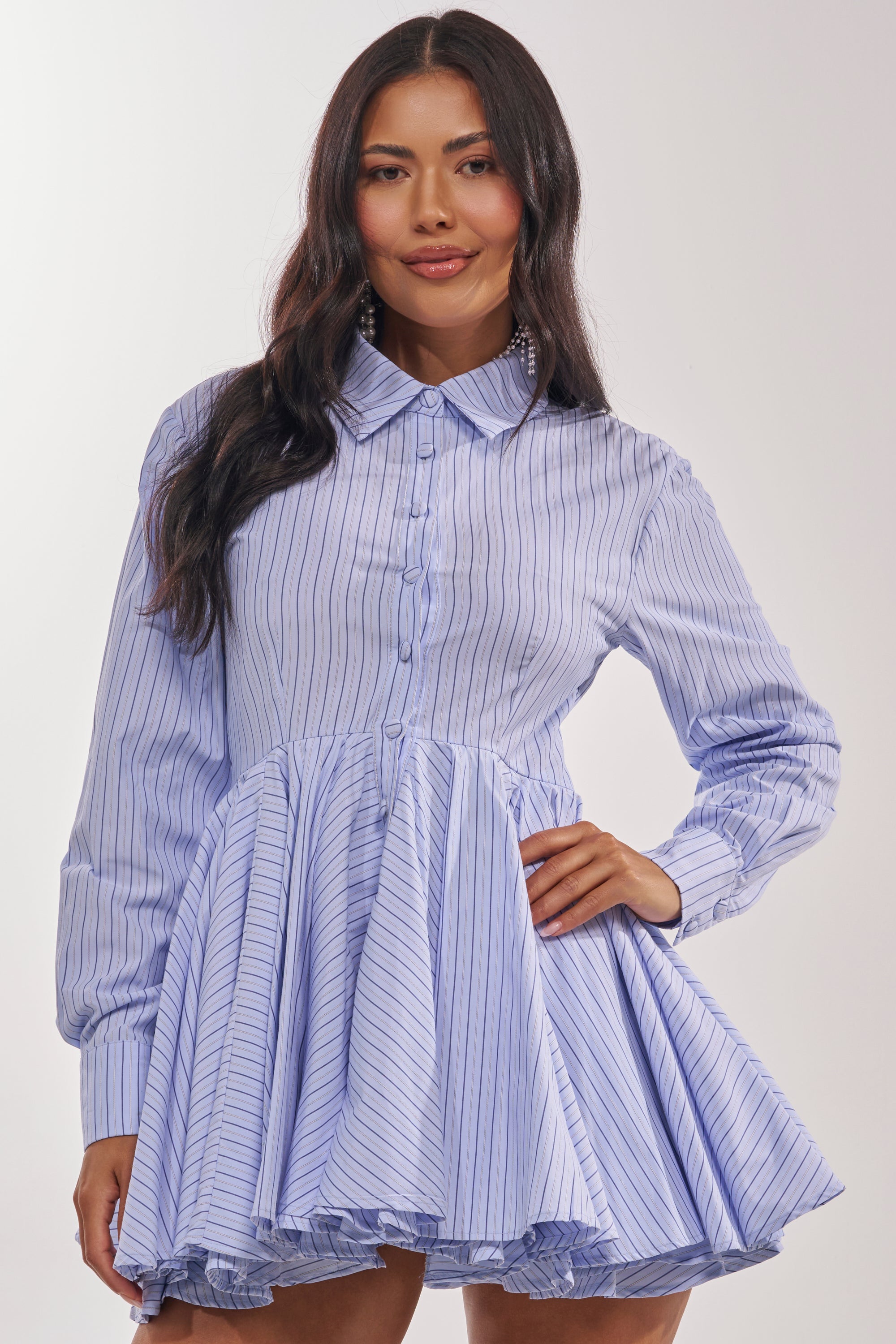 A woman poses confidently in the HAPPY HOUR PINSTRIPE MINI DRESS, featuring blue-and-white poplin pinstripes, long sleeves, a classic collar, and a flared short skirt—perfect for transitioning from office to happy hour. 3