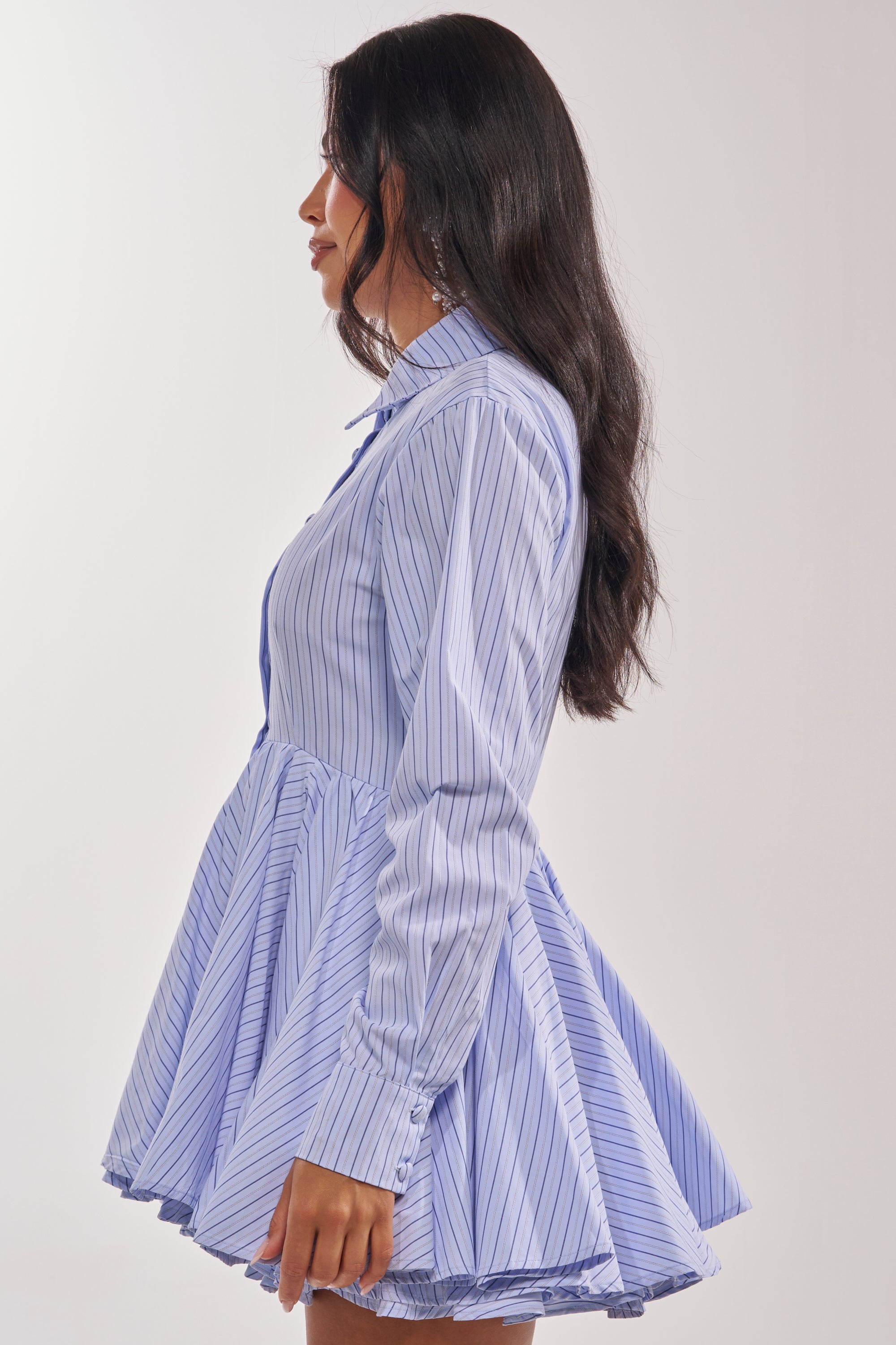 A woman with long dark hair stands in profile, wearing the HAPPY HOUR PINSTRIPE MINI DRESS—featuring blue-and-white stripes, button details, and a flared skirt—perfect for stylish transitions from office to happy hour. 4