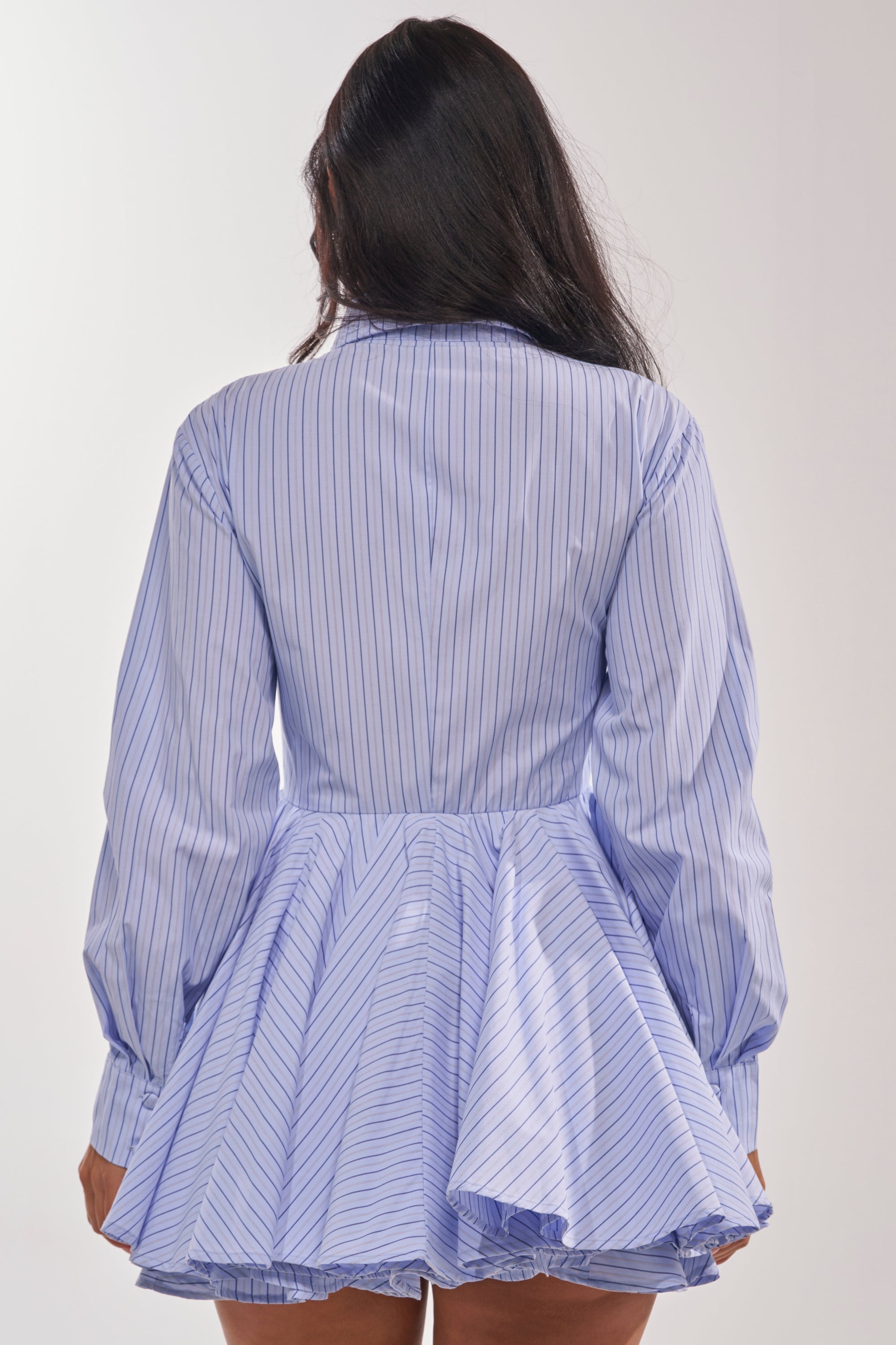A woman with long dark hair faces away, wearing the HAPPY HOUR PINSTRIPE MINI DRESS—a blue-and-white cotton poplin mini dress featuring pinstripes, long sleeves, and a flared ruffled skirt. The background is plain and light-colored. 5