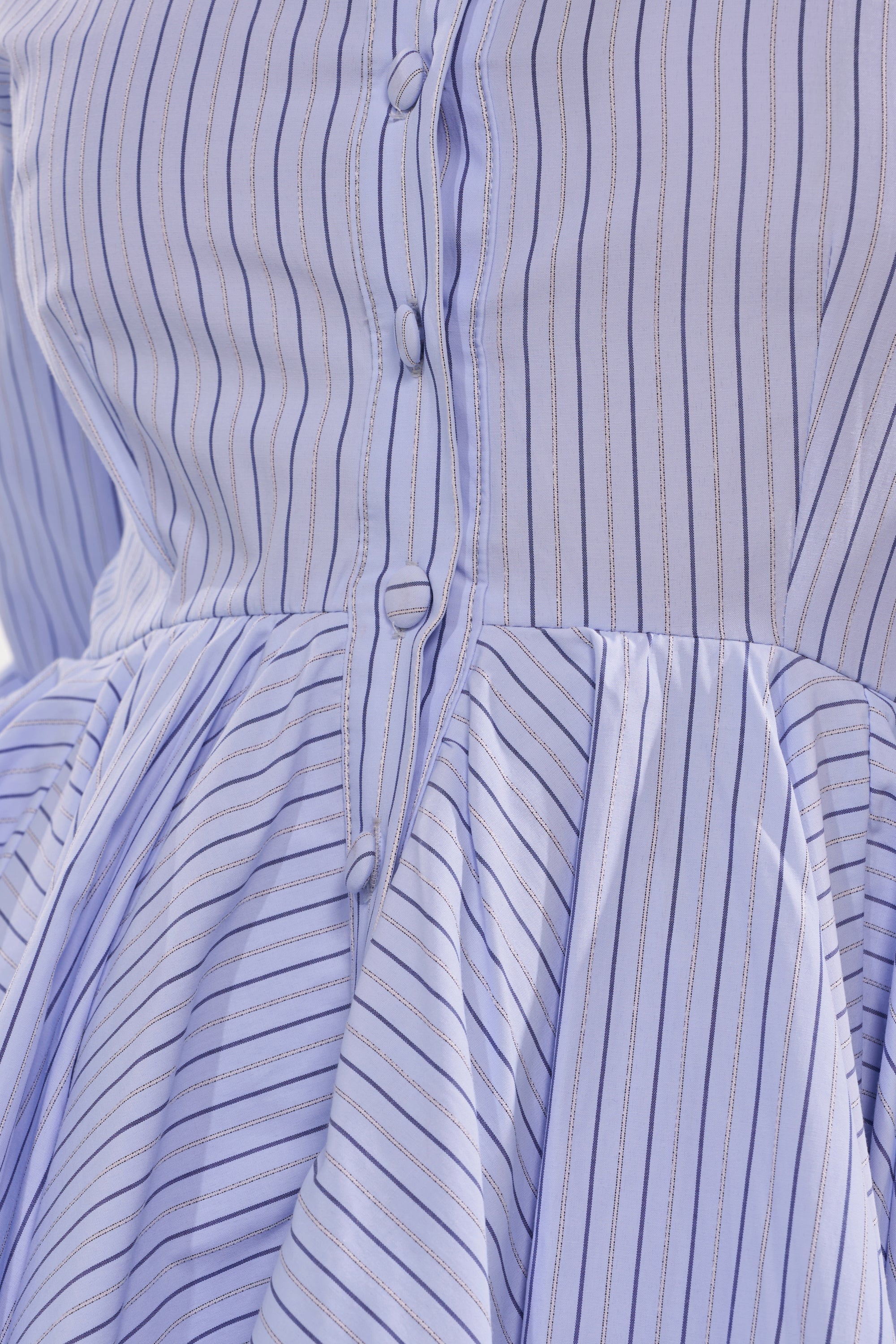 Close-up of the HAPPY HOUR PINSTRIPE MINI DRESS in blue-and-white cotton poplin, featuring fabric-covered front buttons, a fitted bodice, angled stripes, and a gathered skirt—perfect to transition from office to happy hour. 9