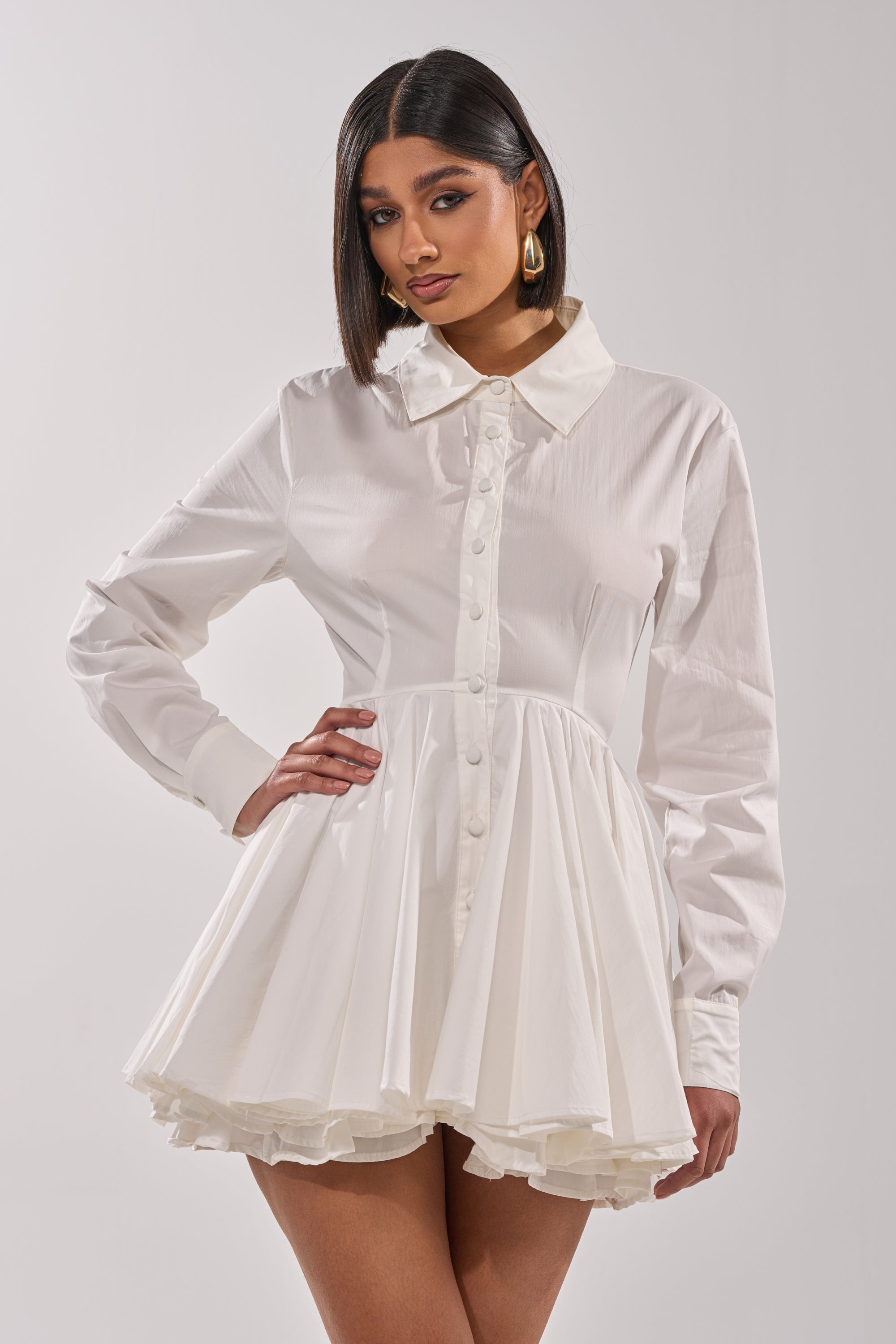 A woman with straight dark hair models THE IT GIRL POPLIN MINI DRESS, a white mini dress with long sleeves and a flared, pleated skirt. She poses with one hand on her hip against a plain light background.