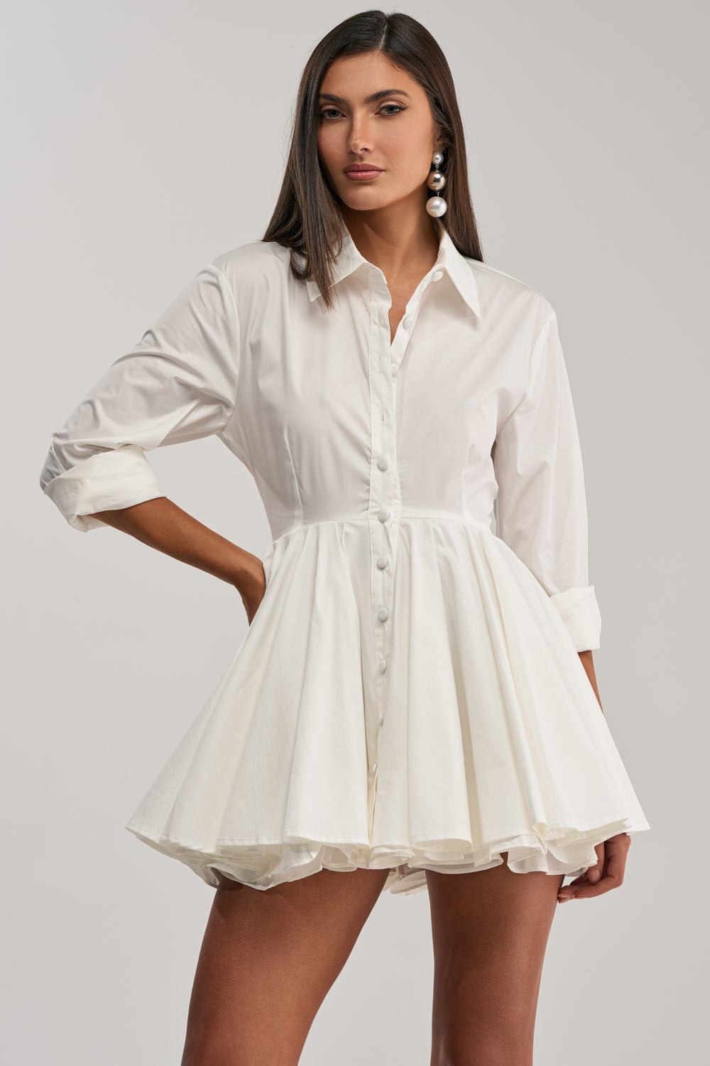 A woman models THE IT GIRL POPLIN MINI DRESS—a white mini dress with a fitted waist and flared, ruffled skirt—against a light gray background. She accessorizes with large pearl earrings for a chic office-to-cocktail look.