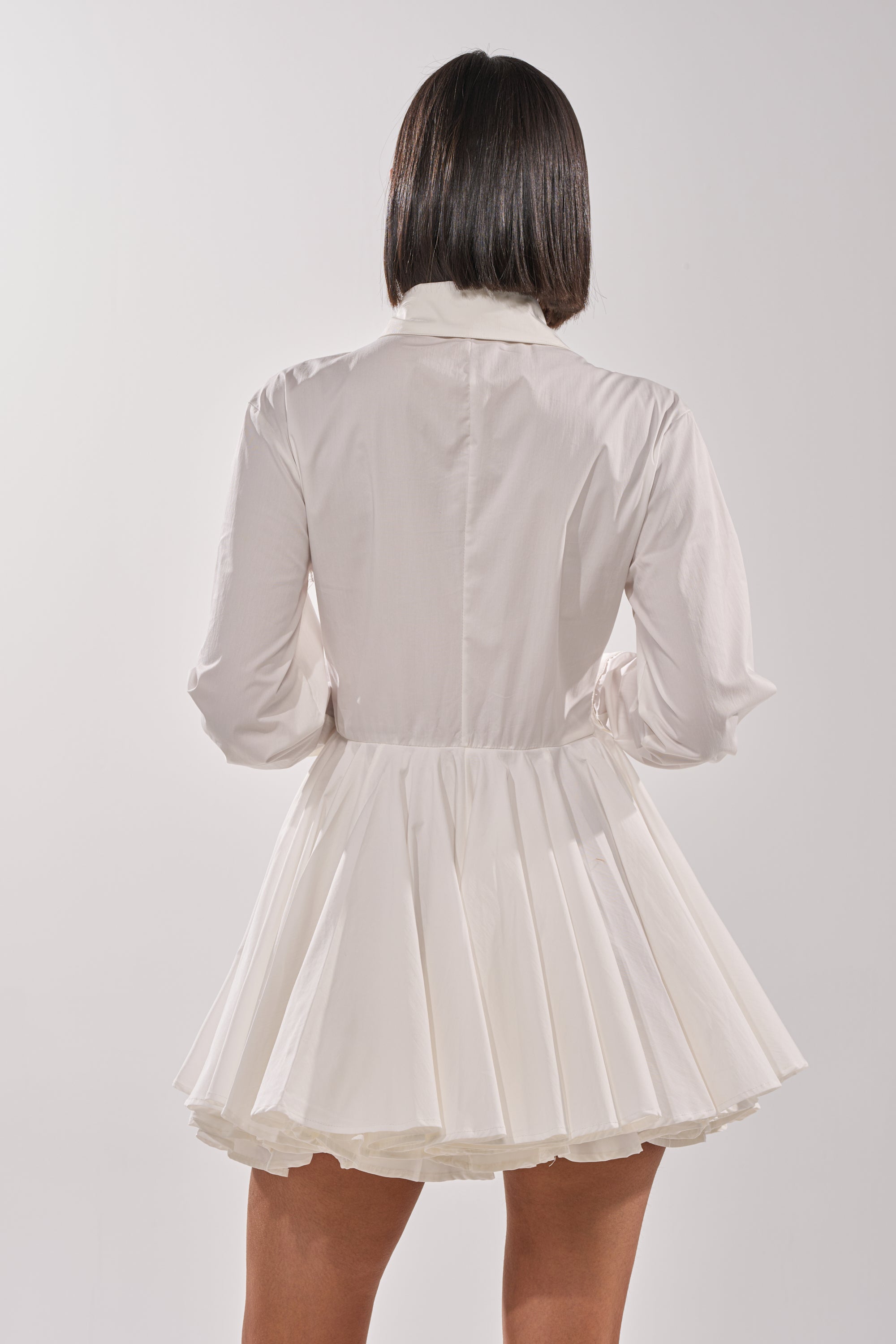 A person with straight, shoulder-length hair stands with their back to the camera against a plain light background, wearing THE IT GIRL POPLIN MINI DRESS in white with long sleeves. 2