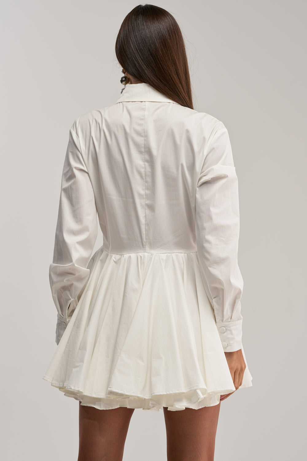 A woman with long brown hair, shown from the back, wears THE IT GIRL POPLIN MINI DRESS—a white mini dress with long sleeves, collared neckline, and flared skirt—styled elegantly against a plain light background.