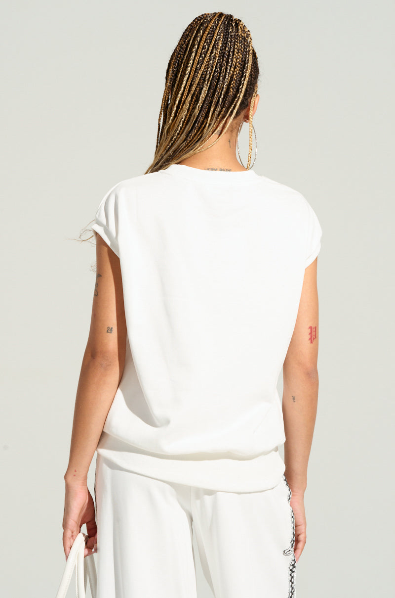 A person with long braided hair, wearing a DANCING IN THE DARK FRENCH TERRY TSHIRT and matching pants, is standing with their back to the camera, holding a white bag in their left hand. Adorned with large hoop earrings and tattoos on their arms, they exude a moto-inspired look straight from the AKIRA Label Dancing In The Dark collection.