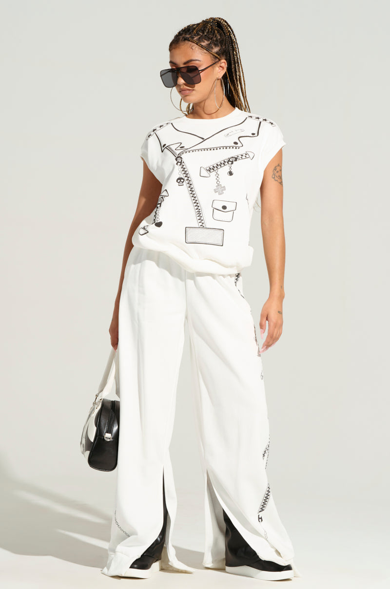 A person with long braided hair wearing oversized black sunglasses, a white DANCING IN THE DARK FRENCH TERRY TSHIRT, and wide-leg white pants poses against a plain background. Holding a small black and white handbag, their stance exudes confidence and style reminiscent of the AKIRA Label.