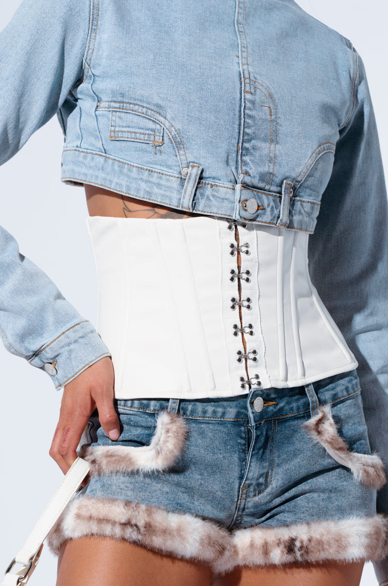 Person wearing a cropped denim jacket, a white corset with hook and eye closure, and high-waisted denim shorts trimmed with fur. Their left hand rests near their pocket, and their right hand holds the CINCHED UP PU BELT by the white strap. The background is a light, neutral color.