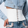 Person wearing a cropped denim jacket, a white corset with hook and eye closure, and high-waisted denim shorts trimmed with fur. Their left hand rests near their pocket, and their right hand holds the CINCHED UP PU BELT by the white strap. The background is a light, neutral color.