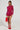 A person with braided hair stands confidently against a white background, wearing the AKIRA Label "If Looks Could Kill" Rhinestone Mini Dress in Red and holding a red feathered bag. The curve-hugging silhouette paired with red high heels creates a bold, stylish look.