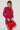 A person in an IF LOOKS COULD KILL RHINESTONE MINI DRESS IN RED, featuring a curve-hugging silhouette, holds a red handbag adorned with feathers. Their long, braided hair and small tattoos are visible on their hands as they stand against a plain white background.
