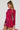 A person with braided hair wears the "If Looks Could Kill Rhinestone Mini Dress in Red," which features an exposed back and long sleeves. The dress's short hemline complements the small red accessory they hold while he or she looks to the side against a plain background.