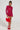 A person stands against a white background, showcasing the IF LOOKS COULD KILL RHINESTONE MINI DRESS IN RED paired with matching red heels. They carry a red feathered handbag and sport long braided hair, with the dress's exposed back enhancing their elegant appearance.