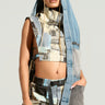 A woman with braided hair poses wearing the PLAY WITH ME DENIM HOOD, a sleeveless patchwork denim crop top paired with matching pants from the AKIRA Label. Her oversized hood is up, revealing tattoos on her arm against a plain neutral background.