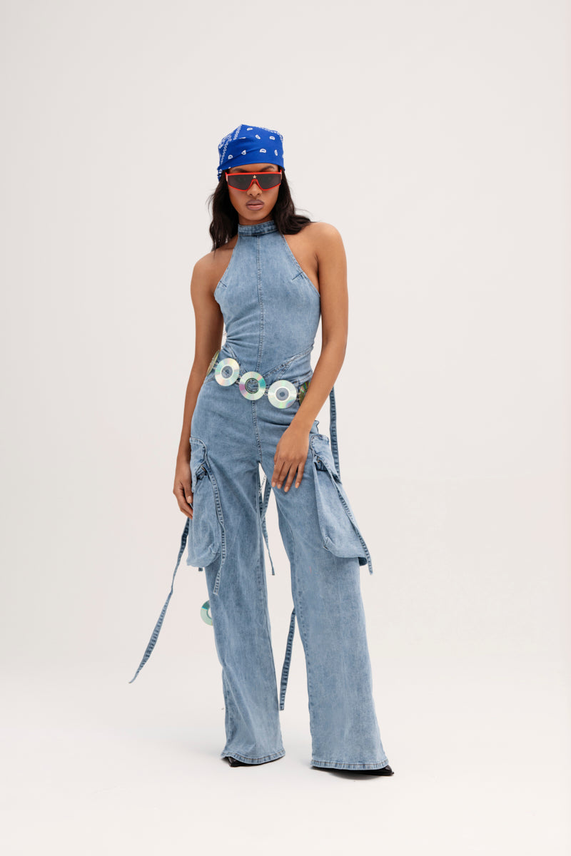 A person radiates confidence in the DON'T YOU DARE DENIM JUMPSUIT IN BLUE, featuring a light-wash halter-neck design with matching wide-leg pants. They complete the look with a blue bandana adorned with white patterns, red sunglasses, and a striking circular-decorated belt against a plain white backdrop. 0