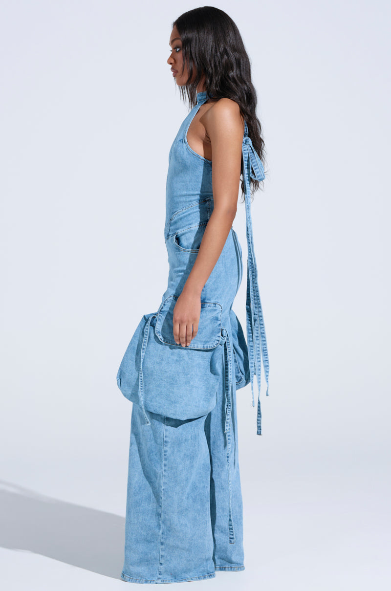 Against a light background, a person with long dark hair stands in profile wearing the DON'T YOU DARE denim jumpsuit in light blue, featuring wide-legged pants with oversized pockets. They carry a matching denim backpack over one shoulder, accentuating the outfit's wide-leg silhouette. 4