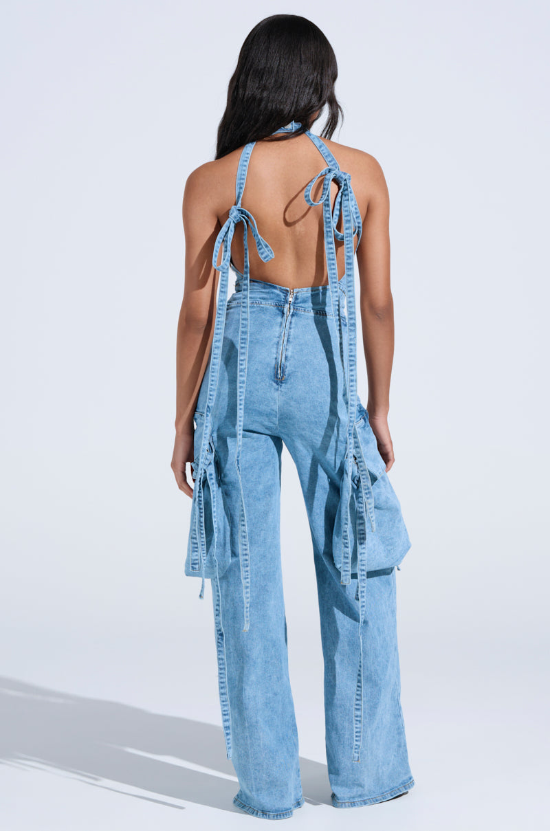 A person with long hair sports the DON'T YOU DARE DENIM JUMPSUIT IN BLUE, featuring a backless light-wash halter top and high-waisted wide-leg jeans. The ensemble highlights large side pockets and decorative ties against a plain white backdrop. 5