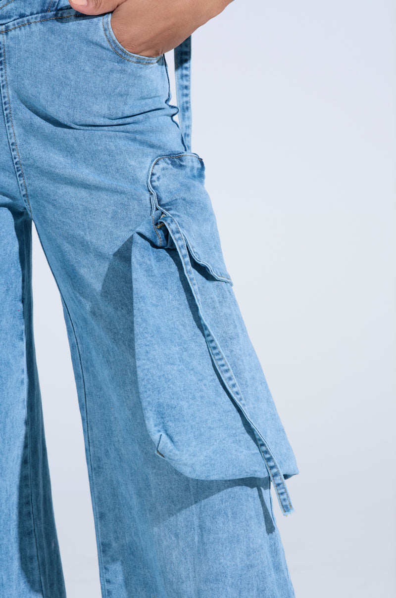 Close-up of a person wearing the DON'T YOU DARE Denim Jumpsuit in Blue with a light-wash, wide-leg silhouette featuring large flap pockets. Their hand rests in one pocket against an off-white background, enhancing the outfit's look. 8