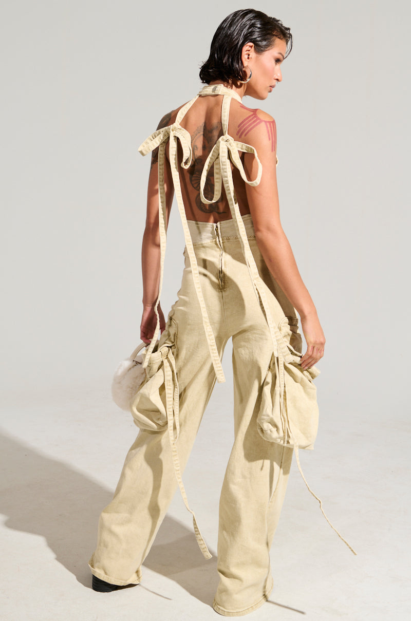 A person with short dark hair stands in the "DON'T YOU DARE DENIM JUMPSUIT," a beige ensemble adorned with bows and wide-legged pants, complemented by cargo-style pockets. They hold a small, fuzzy white bag, revealing a large tattoo on their open back. The background maintains a neutral tone.