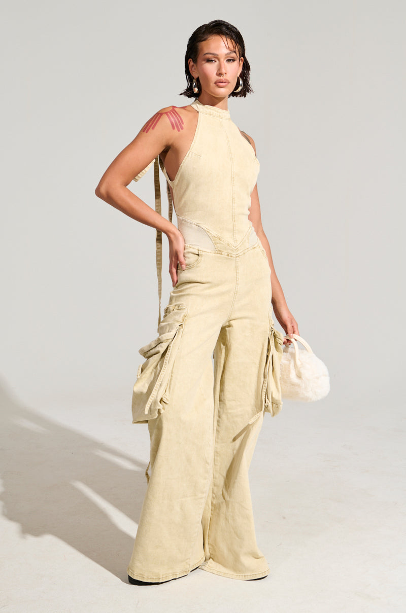 Wearing the DON'T YOU DARE DENIM JUMPSUIT, a person with short hair confidently strikes a pose. The chic beige jumpsuit showcases cargo-style pockets and an open back. They accessorize with a white handbag and have applied subtle makeup, standing against a plain gray background.
