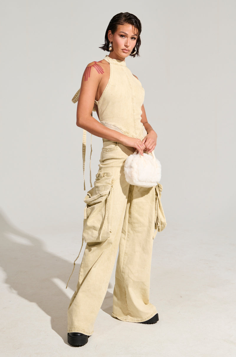 A person stands confidently in the DON'T YOU DARE DENIM JUMPSUIT, featuring a light beige tone with cargo-style pockets and wide-legged pants. They hold a fluffy white handbag and wear large black shoes. The background is a plain white studio setting.
