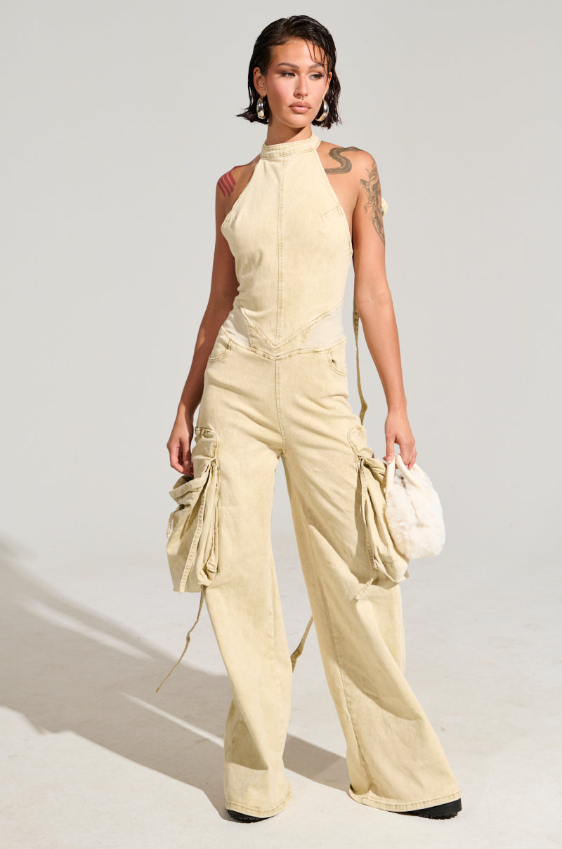 A person stands confidently in a DON'T YOU DARE DENIM JUMPSUIT, showcasing an open back, halter neck, and wide legs with ruched side details. They hold a small, furry white bag and have tattoos on one arm against a simple light gray background.