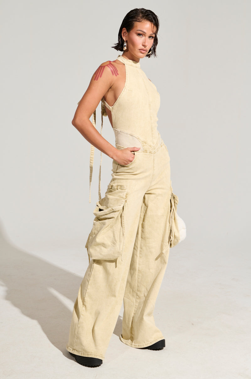 A person with short, wet-look hair poses confidently in a sleeveless DON'T YOU DARE DENIM JUMPSUIT featuring wide legs and cargo-style pockets. Their hands are tucked casually in their pockets, while several red streaks accent their shoulders against a plain gray background.