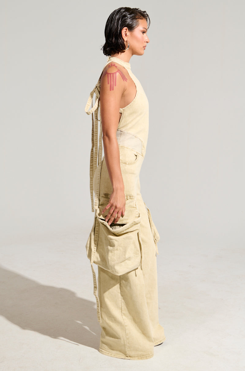 A person stands in profile against a plain background, wearing the DON'T YOU DARE DENIM JUMPSUIT, featuring sleeveless tan fabric with cargo-style pockets and a tied back detail. Their short, wet-look hair complements the pink, drip-like design on their upper arm.