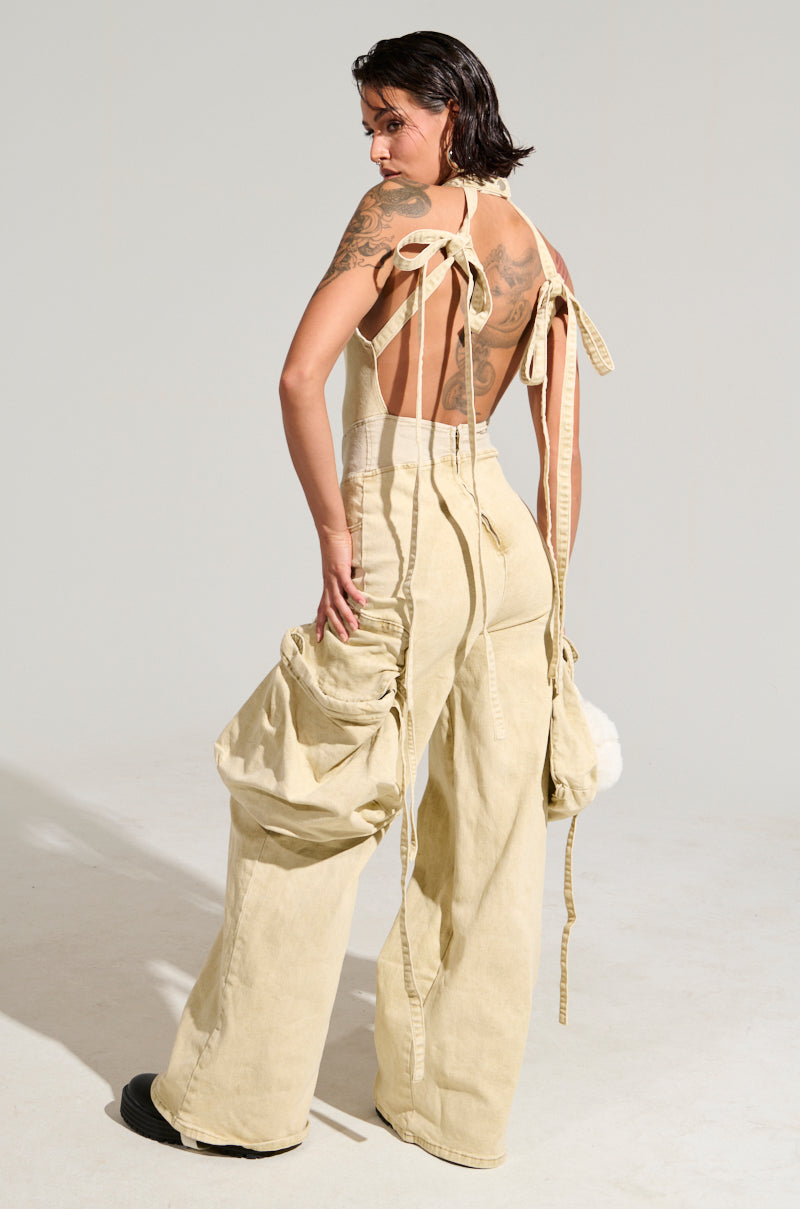 A person with tattoos on their back is showcasing the DON'T YOU DARE DENIM JUMPSUIT, which features an open-back design and wide-leg silhouette complete with large cargo-style pockets. The outfit includes loose straps that drape down, and they are posing against a plain background.