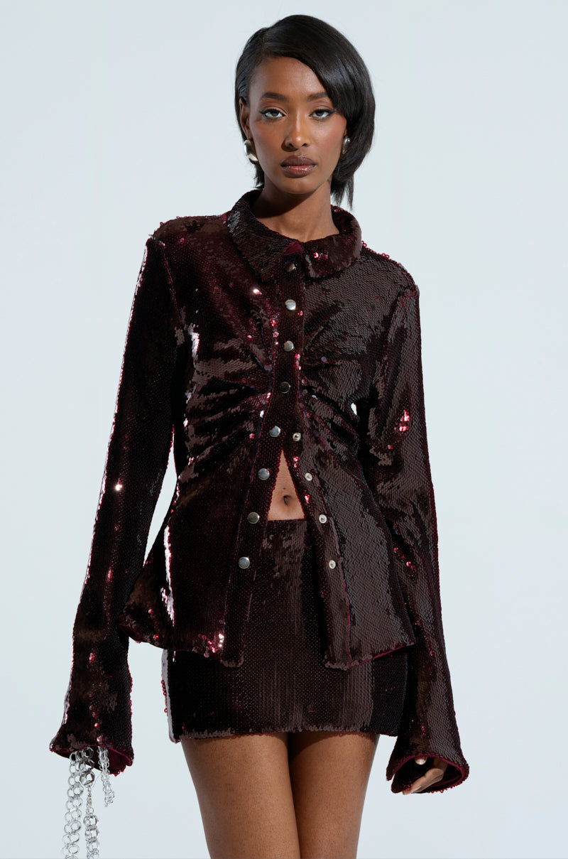 A woman showcases the LIGHTS OUT BUTTON DOWN BLOUSE, a dark sequined blouse featuring long sleeves and paired with a coordinating skirt. Her hair is elegantly styled to one side as she holds a silver chain in her left hand against a simple backdrop, while understated ruching enhances the sophistication of her look.