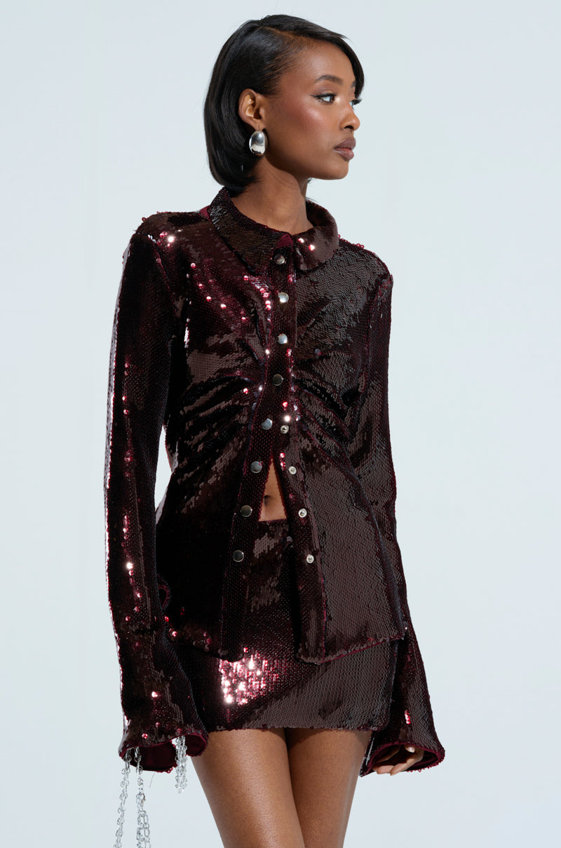 A woman captivates in the LIGHTS OUT BUTTON DOWN BLOUSE with shimmering dark sequins and ruched detailing, complete with long sleeves and a short skirt. Her sleek hair perfectly complements her silver hoop earrings as she gazes to the side against a plain, light-colored backdrop, exuding elegance and style.