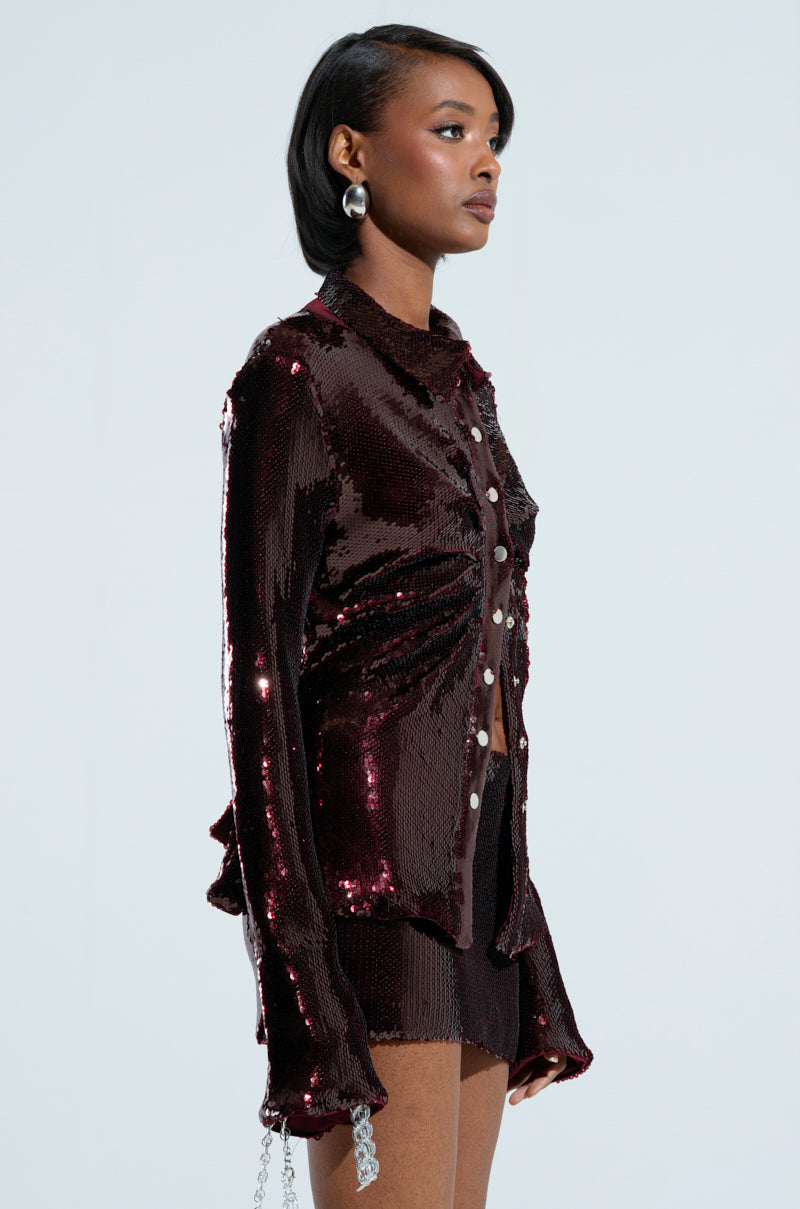 A woman captures attention with a side profile view, showcasing elegance in her dazzling burgundy **LIGHTS OUT BUTTON DOWN BLOUSE** paired with coordinating shorts. The ruched accents enhance her chic short black hair and oversized silver earrings, culminating in a fashionable and captivating ensemble.
